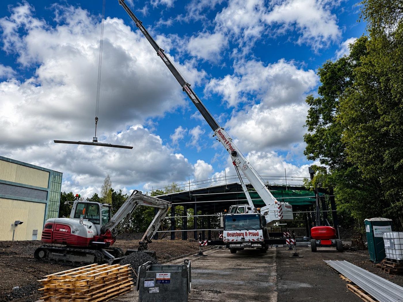 A crane lifting a beam beside the part-built steel frame, with an excavator in the foreground